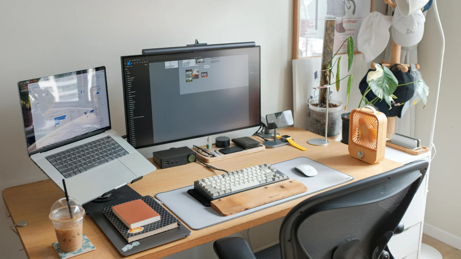 Home office desk with a laptop, monitor, keyboard, notebooks, and iced coffee beside a window.